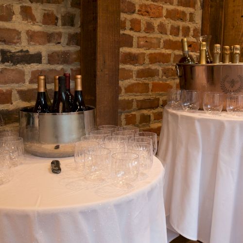 Tables with wine bottles in buckets, surrounded by empty glasses, set against a brick wall.