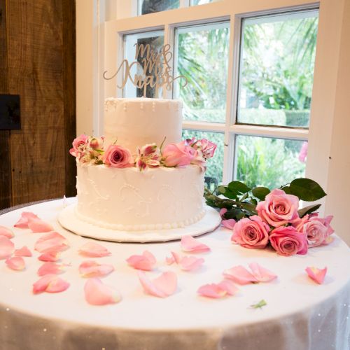A white two-tiered cake with a floral topper is surrounded by pink roses and petals on a table in a bright room.