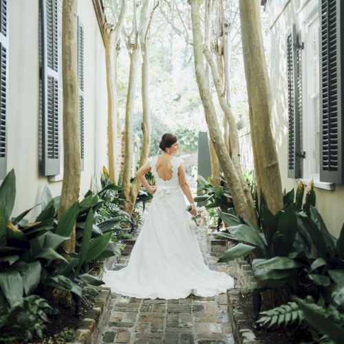 A bride in a white dress stands in a narrow, tree-lined path with plants and shutters on both sides, looking away from the camera.