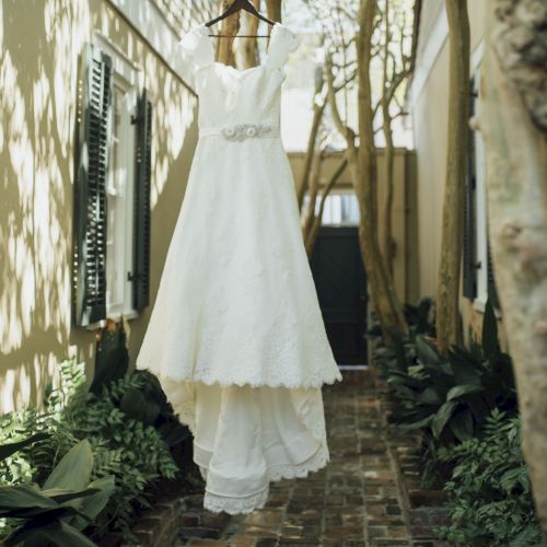 A white wedding dress hangs from a branch in a garden-lined walkway with brick pavement and shuttered windows.