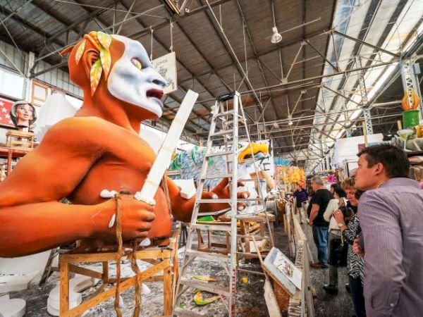 A craftsman wearing a large orange mask sculpts a figure with a white head, holding a tool in a workshop filled with sculptures and onlookers.