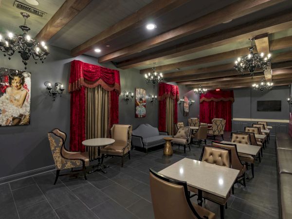 A stylish lounge or cafe with red curtains, chandeliers, and velvet seating; small square tables and cushioned chairs line dark walls.