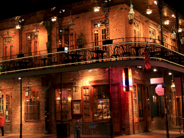 A brick building with a corner caf&eacute;, two stories of balconies, warm lights, lanterns, and pedestrians at night, cozy urban street scene.