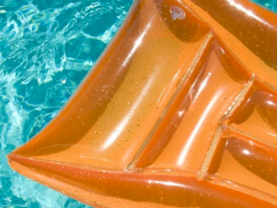 A close-up of an orange inflatable boat floating on turquoise water, showing its glossy, ribbed sections and sunlit reflections.