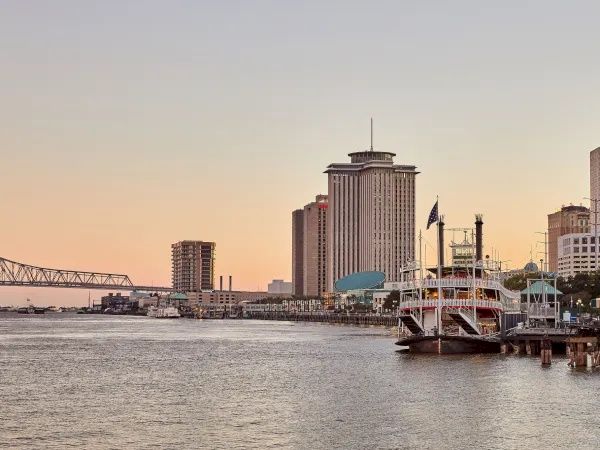 A waterfront city skyline at sunset, with a tall tower and a docked boat along the riverbank, under a pastel sky.