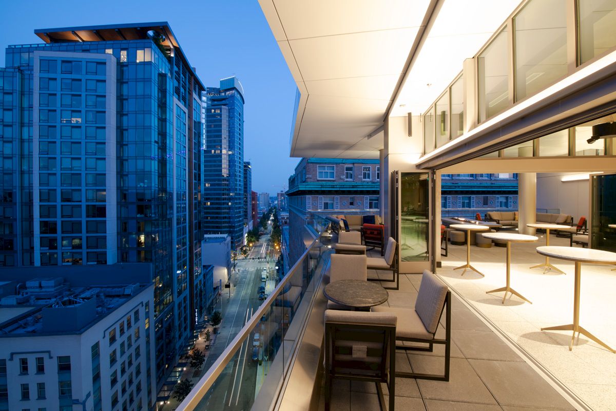 A cityscape at dusk with a modern balcony, seating, tables, and tall buildings.