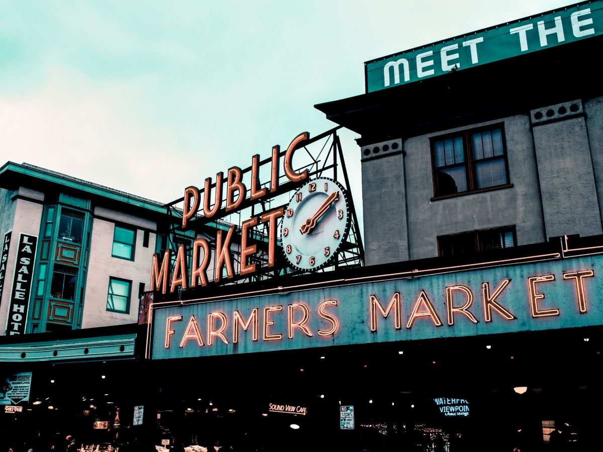A vintage-style sign for a public farmers market with a clock, set against an urban backdrop with building facades.