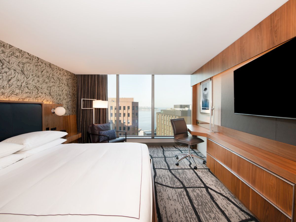 A modern hotel room with a large bed, desk, TV, and a view of buildings through a window.
