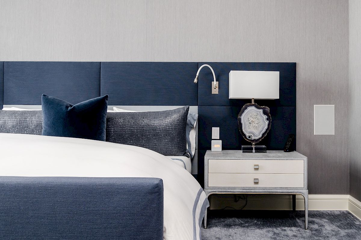 A modern bedroom with a blue headboard, pillows, a white nightstand with a lamp, and a decorative mirror.