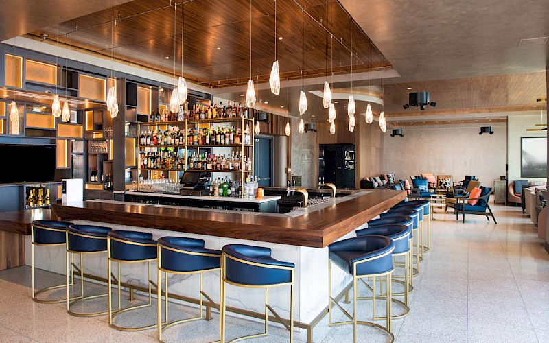 The image shows a modern bar with a wooden counter, blue cushioned stools, hanging lights, and a well-stocked liquor shelf in a stylish setting.